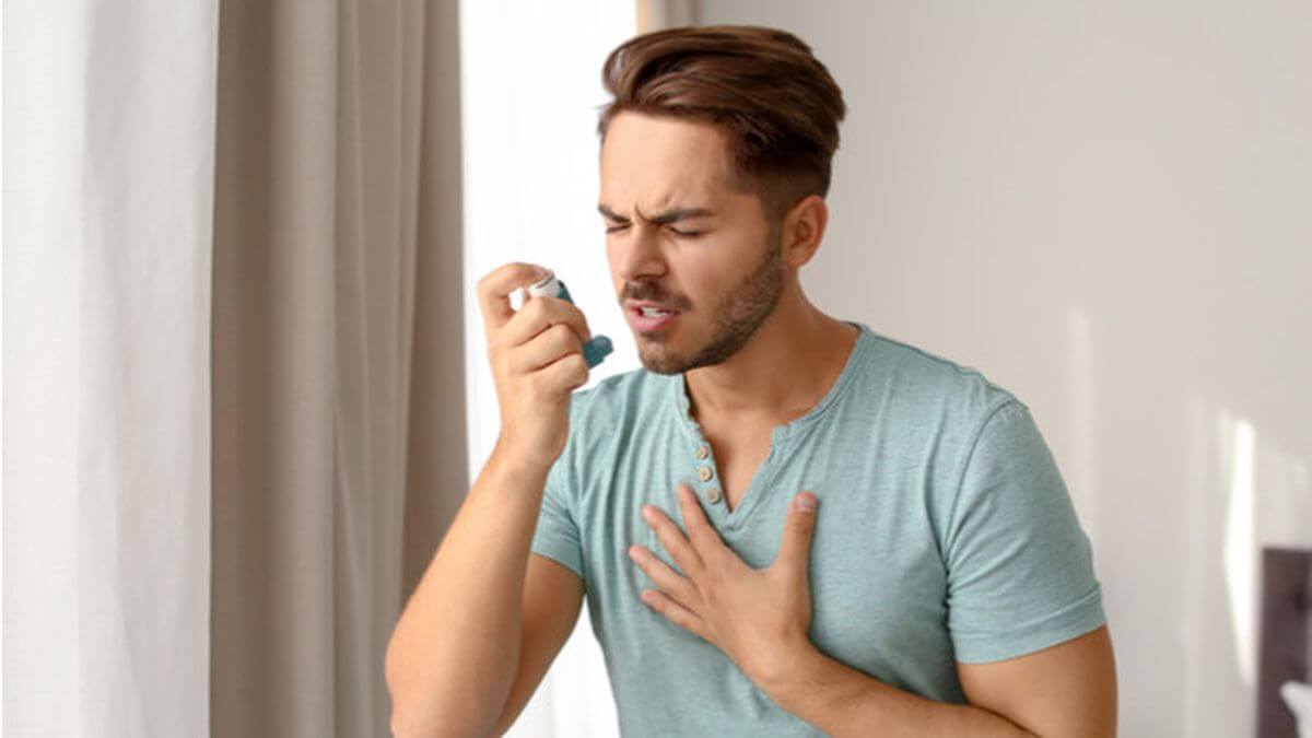 Young man using a blue asthma inhaler with hand on chest, experiencing an asthma attack at home