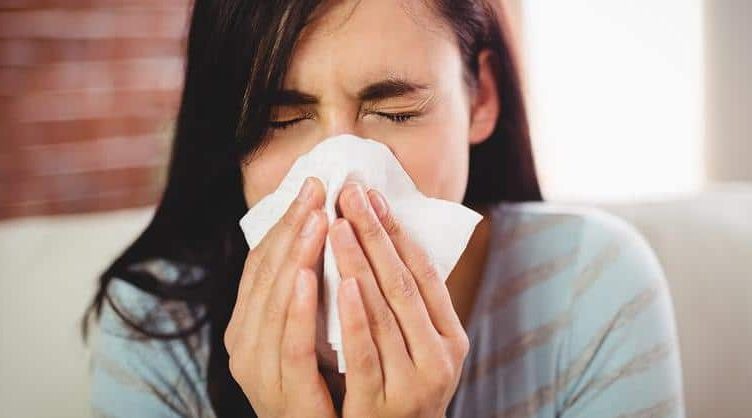 Woman blowing her nose into a tissue with eyes closed, showing cold and flu symptoms at home