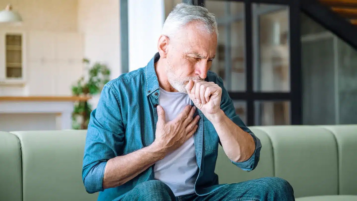 Older man sitting on a sofa coughing into his fist with hand on chest, showing bronchitis symptoms